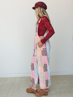 A side-profile shot of a model in a wide-leg patchwork floral jumpsuit layered over a red crochet long-sleeve top, styled with a burgundy baker boy cap and tan studded clogs — the whole look has a nostalgic, quilt-meets-70s energy that feels both pla
