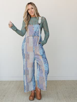 A wide-leg patchwork floral overall jumpsuit in soft denim tones styled over a sage lace long-sleeve top, paired with tan clogs — the kind of outfit that somehow looks effortless and thought-out at the same time.