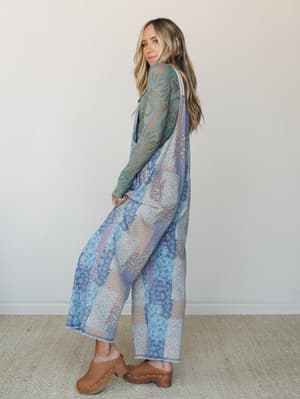 A model shot over the shoulder in a wide-leg patchwork floral jumpsuit layered over a sage lace long-sleeve top, styled with tan wooden clogs — the whole look feels like something you'd find in a vintage shop and never want to return.