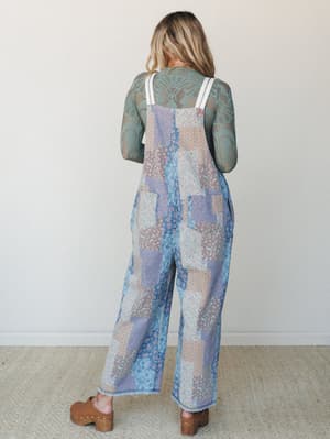 A back-facing model wears wide-leg patchwork floral overalls in soft denim tones layered over a sage green lace long-sleeve top, styled with brown wood clogs — the kind of outfit that looks like it was put together effortlessly but absolutely wasn't.
