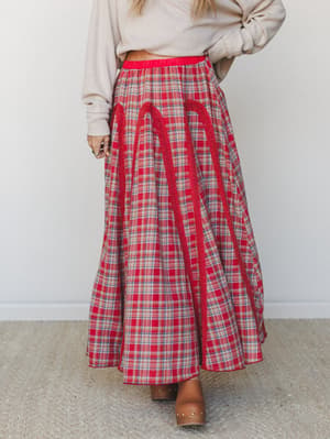 A full, sweeping red plaid maxi skirt with lace trim detailing and an elastic waistband, styled with a knotted cream sweater and brown clogs for an effortless Western-boho feel.