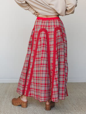 A red plaid maxi skirt shot from the back, styled with a cropped oatmeal sweater and wood-platform clogs — the lace arch appliqué details and full gathered silhouette give it that sweet spot between vintage and boho.