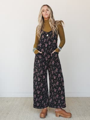 A flowy wide-leg floral jumpsuit layered over an olive turtleneck, styled with wood-sole clogs and stacked turquoise bracelets — the kind of effortless fall outfit that looks like it came together on purpose.