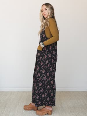A floral maxi jumpsuit in black with scattered pink blossoms is layered over a mustard long-sleeve turtleneck, styled with tan wood-platform clogs and a stack of beaded bracelets — the kind of outfit that looks like it came together effortlessly but
