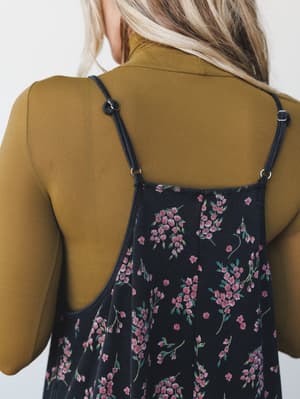 A close-up back shot of a floral spaghetti-strap jumpsuit layered over an olive turtleneck long-sleeve — a cool-weather styling moment that feels both vintage and effortless.