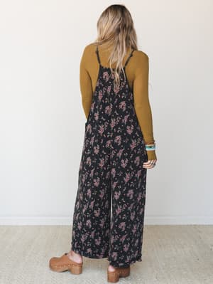 A back-view shot of a wide-leg floral jumpsuit in deep black with scattered pink floral sprigs, layered over a mustard long-sleeve top and styled with tan wooden clogs and stacked bracelets — the kind of outfit that looks effortless but is clearly ve
