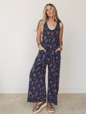A wide-leg floral jumpsuit in deep navy with tiny pink ditsy blooms — worn with layered necklaces, stacked bracelets, and platform sandals for a look that feels effortlessly put-together without trying too hard.
