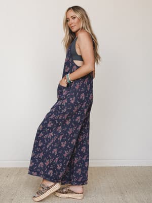 A model shot in three-quarter profile showing a wide-leg floral jumpsuit in deep navy with ditsy pink floral print, styled with a gray bralette underneath, layered bracelets, and leopard print platform espadrilles — relaxed and a little romantic.