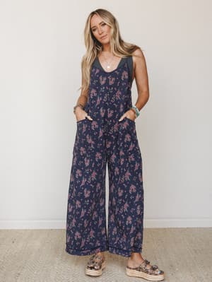 A wide-leg floral jumpsuit in deep navy with scattered pink ditsy blooms, styled with platform espadrille sandals, stacked bracelets, and a delicate pendant necklace — effortlessly put-together without trying too hard.