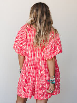 A back-view shot of a cherry red gingham mini dress with voluminous puff sleeves and white eyelet trim detailing, styled with a stack of beaded and turquoise bracelets against a soft neutral studio backdrop.