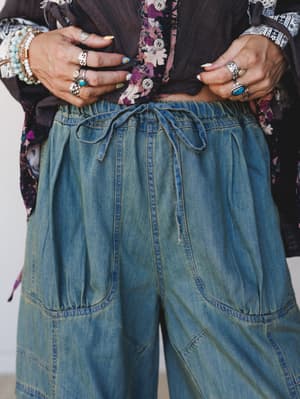 A close-up detail shot showing wide-leg denim cargo pants in a worn, faded wash, styled with a dark gauze top and floral kimono — the stacked silver rings and turquoise jewelry doing exactly as much work as the outfit.
