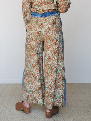 A back-view shot of wide-leg paisley pants with denim side panel detailing and a frilled denim waistband, styled as a matching set with a coordinating top and paired with wood-studded clogs — earthy, expressive, and very much a whole mood.