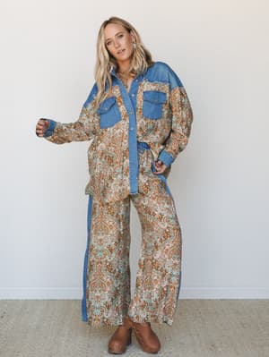 A matching paisley set styled with denim colorblocking — wide-leg pants and an oversized button-down shirt share the same warm brown and turquoise print, grounded by tan clogs and a relaxed, lived-in confidence.