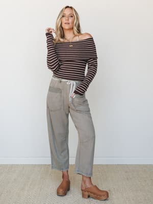 A relaxed full-body shot of a model in an off-shoulder striped sweater tucked into wide-leg barrel pants in ash olive, paired with tan platform clogs — the kind of effortless, earthy outfit that feels as good as it looks.