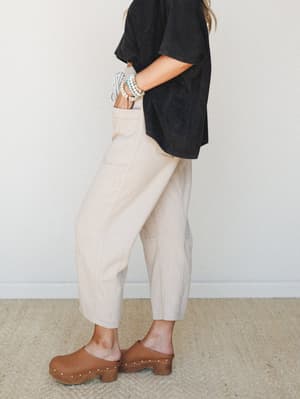 A side-profile shot of cropped barrel-leg pants in a warm oatmeal herringbone, styled effortlessly with a faded black oversized tee, wood-soled clogs, and stacked bead bracelets — the kind of outfit that looks like it came together without trying.