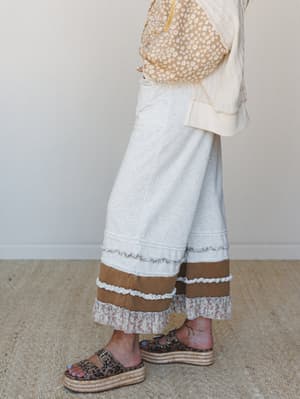 A side-angle mid-shot focused on wide-leg pants in light heather gray with layered ruffle hem detailing in tan and floral fabric, styled with a daisy-print top and leopard platform espadrille sandals against a soft neutral interior wall.