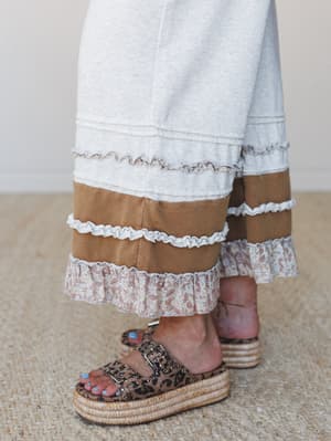 A close-up of wide-leg pants in light heather gray with mixed-fabric tiered ruffle trim at the hem — part cozy sweater knit, part floral cotton, part suede-like camel panel — styled with leopard print platform sandals on a jute rug.