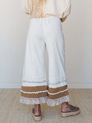 A back-view shot of wide-leg heather gray pants with layered ruffle and colorblock hem detailing, styled with a floral top, cream knit, and platform espadrilles — the kind of outfit that looks effortless but clearly had a point of view.