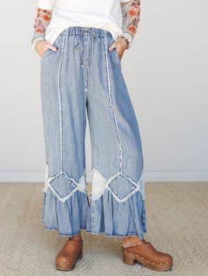 Wide-leg denim pants with ruffled hems and frayed patchwork detailing styled with a white tee, floral mesh sleeves, and tan clogs — the kind of outfit that looks effortless but was absolutely thought about.