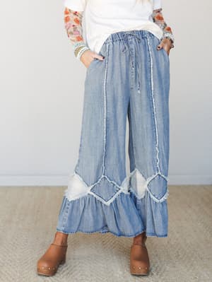 Wide-leg denim pants with ruffled hems and raw-edge diamond patchwork detailing styled with a white top, floral mesh sleeves, and tan clogs — effortlessly boho and full of personality.