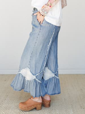 A side-profile shot of wide-leg denim pants with dramatic ruffled hems and frayed patchwork detailing, styled with a floral embroidered sheer-sleeve top and tan wooden clogs — the kind of outfit that makes a Tuesday feel like a road trip.