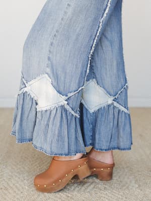 A close-up of wide-leg denim flares with ruffled hems and raw-edge patchwork detail, styled with tan studded clogs on a jute rug — the kind of jeans that make a regular Tuesday feel like a road trip.