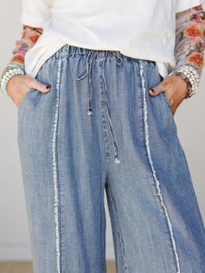 A close-up shot of relaxed wide-leg denim pants with exposed frayed seams and a drawstring elastic waist, styled with a white tee and a floral-sleeved layer — hands tucked in pockets, stacked bracelets on both wrists, very much a 'I look this good on
