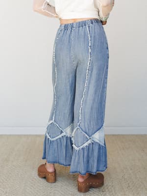 A back-view shot of wide-leg denim pants with ruffle hem and exposed frayed seam detailing, styled with a sheer cream top and wood-platform clogs — the kind of outfit that makes a regular Tuesday feel more intentional.