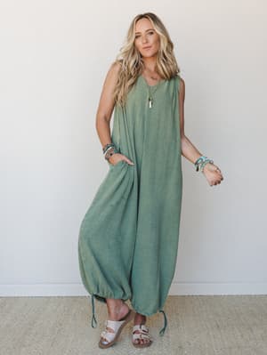 A flowy, wide-leg jumpsuit in a soft juniper green gauze fabric — relaxed and roomy with elasticized ankles and side pockets, styled with layered boho bracelets and flat sandals for that effortless, 'I just threw this on and somehow look amazing' ene