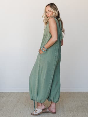 A breezy sage green gauze jumpsuit with balloon-hem legs and a shoulder tie closure, shot from a three-quarter back angle against a soft white studio wall — relaxed and effortless in the best possible way.