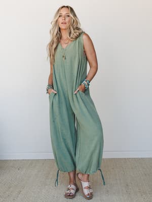 A relaxed, full-body shot of a model in a sage green sleeveless gauze jumpsuit with balloon-style cropped legs, styled with beaded bracelets, a layered pendant necklace, and white buckle sandals against a clean white wall — effortless and earthy in t