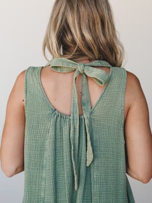 A back-detail shot of a sage green gauze dress with a soft tie bow at the nape — the kind of open-back moment that feels effortless rather than trying.