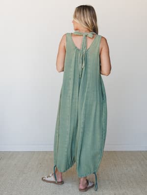 A wide-leg, balloon-silhouette jumpsuit in washed sage green gauze is shown from the back, with a standout open-back tie closure at the neckline and ankle-cinch hem — effortless and a little unexpected.