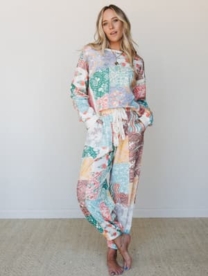 A matching patchwork printed set — relaxed sweatshirt and jogger pants in a kaleidoscope of soft florals, paisleys, and botanical blocks — worn barefoot against a neutral studio wall, feeling effortlessly at-home and quietly expressive.