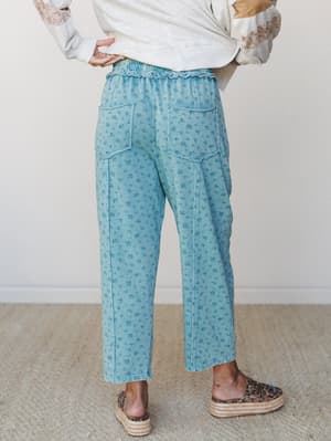 Shot from behind, a model wears relaxed cropped pants in washed teal blue with an allover ditsy floral print, paired with an oatmeal graphic sweatshirt and leopard espadrille flatforms — the kind of outfit that feels effortless but clearly thought th