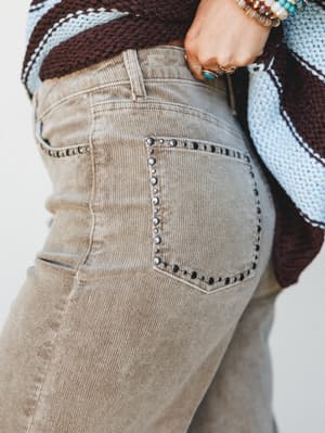 A close-up detail shot of taupe corduroy trousers with studded back pocket trim, styled with a chunky color-block knit sweater and stacked turquoise and beaded bracelets — earthy, expressive, and very much the kind of outfit you'd wear on purpose.