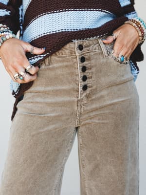 A close-up mid-shot focused on the waist and hip of wide-leg corduroy trousers in a warm tan, styled with a chunky color-block knit sweater and layered silver and stone jewelry — the kind of outfit that looks like it came together effortlessly but de