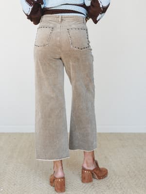 Shot from behind, a model wears wide-leg corduroy pants in a washed sandy tan, styled with a colorblock crochet sweater and studded wood-heel mules — the kind of outfit that makes a Tuesday feel intentional.