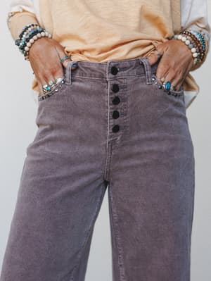 A close-up of wide-leg corduroy trousers in dusty lilac, styled with a peach tee and layered boho jewelry — the kind of pants that make you rethink everything you thought about 'practical.'