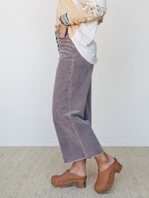 A side-profile shot of wide-leg corduroy trousers in dusty lilac styled with a floral-embroidered top, stacked bracelets, and tan studded clogs — the kind of outfit that feels pulled-together without trying too hard.