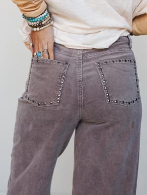 A close-up back view of dusty lilac corduroy wide-leg trousers with studded pocket trim, styled with a cream long-sleeve top and layered boho jewelry — the kind of pants that make a casual outfit feel like a whole personality.