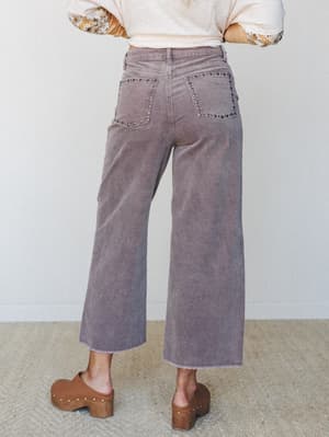 Shot from the back, these wide-leg corduroy trousers in dusty lilac feature studded back pocket trim and a raw hem — styled with a cream top, floral-sleeved layer, and wood platform clogs for an effortlessly earthy, put-together look.