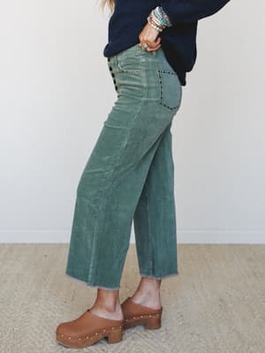 A side-profile shot of wide-leg corduroy crops in a faded juniper green, styled with a navy sweater, wood-heeled clogs, and a wrist full of turquoise bracelets — the kind of outfit that makes a Tuesday feel intentional.