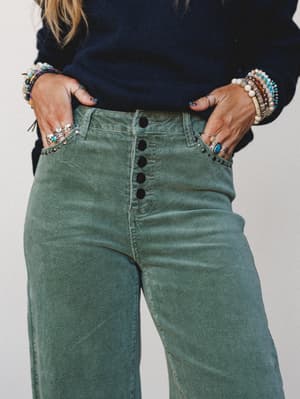 A close-up torso shot of sage green corduroy wide-leg trousers styled with a navy ribbed sweater and an armful of layered beaded and silver bracelets — the kind of outfit that looks like it came together effortlessly but definitely did not.