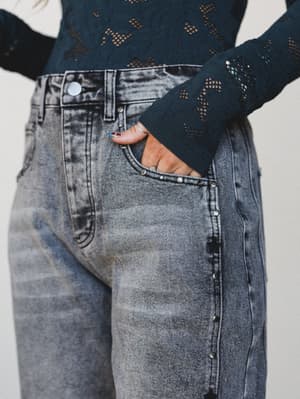 A close-up mid-shot of acid-washed black denim wide-leg pants paired with a teal openwork lace long-sleeve top, hand tucked casually in the front pocket with stacked silver rings visible.