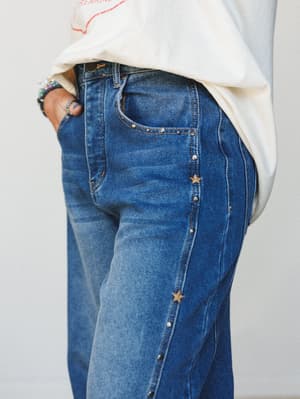A close-up detail shot of medium-wash wide-leg jeans with a side seam lined in alternating star studs and silver rivets, styled with a cream graphic tee and stacked beaded bracelets — the kind of jeans that do the talking for you.