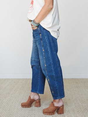 A side-profile shot of wide-leg cropped denim jeans with gold star studs running down the side seam, styled with a graphic tee, stacked bead bracelets, and brown suede platform clogs — easy, expressive, and a little bit rock-and-roll.