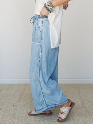 A side-profile shot of wide-leg chambray pants in a soft washed denim, styled with a white tee, beaded bracelets, and buckle sandals — the kind of outfit that feels effortless from morning coffee to wherever the day takes you.