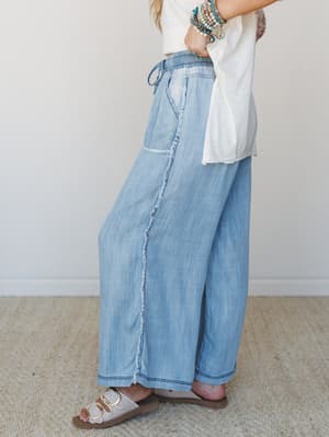 A side-profile shot of wide-leg chambray pants with a frayed seam detail and drawstring elastic waist, styled with a white tee, sandals, and a stacked bracelet arm — easy, relaxed, and just a little effortless.