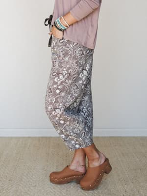 A side-profile shot showing floral barrel-leg crop pants in a mocha and cream print, styled with a dusty mauve top and tan wood-sole clogs — the kind of outfit that feels intentional without trying too hard.